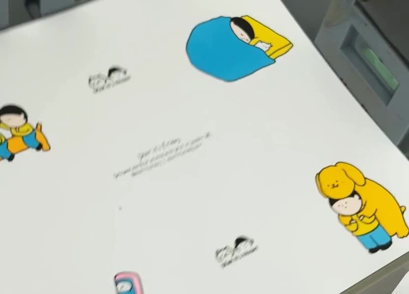 The press running: a two-colour sheet in the tray, a pink sheet coming through, a newsprint poster, and a sheet of blue line drawings.