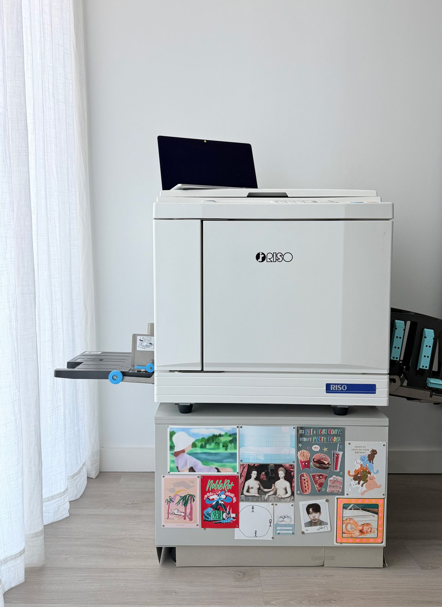The Riso SF9350 in the studio, a white duplicator on a cabinet
                    covered in prints and postcards.